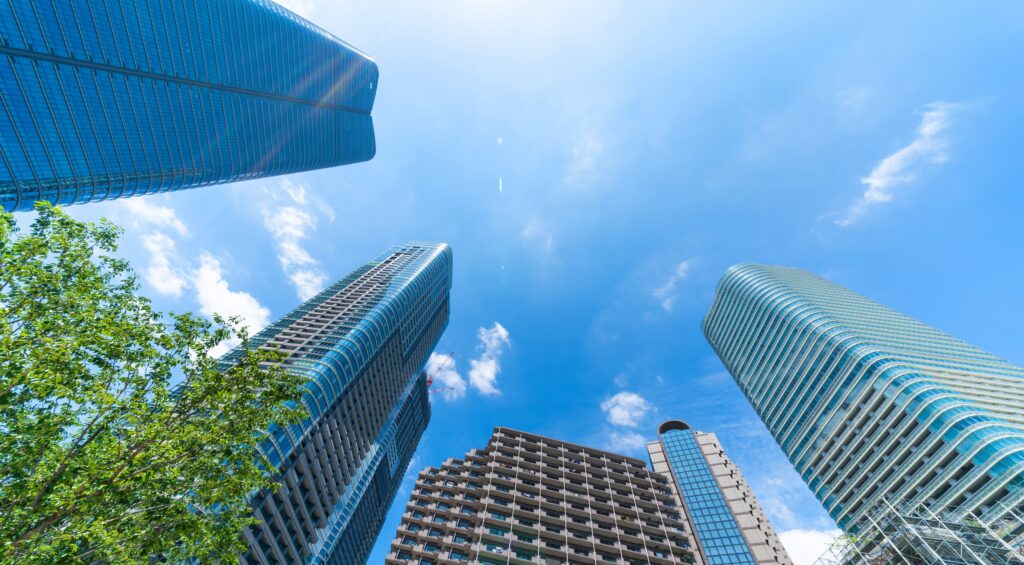 A low-angle shot looking directly up at several modern, tall skyscrapers against a vibrant blue sky, with green tree branches visible in the bottom left corner.
