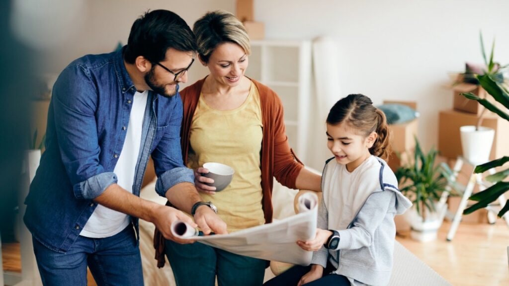 A smiling family of three looking at large, rolled-out blueprints together in a room surrounded by cardboard moving boxes.