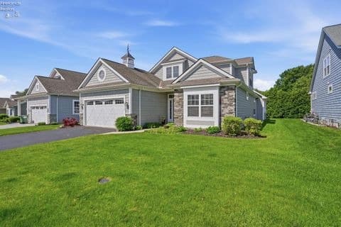A grey suburban home with a white two-car garage, stone facade accents, and a large, bright green front lawn under a sunny blue sky.
