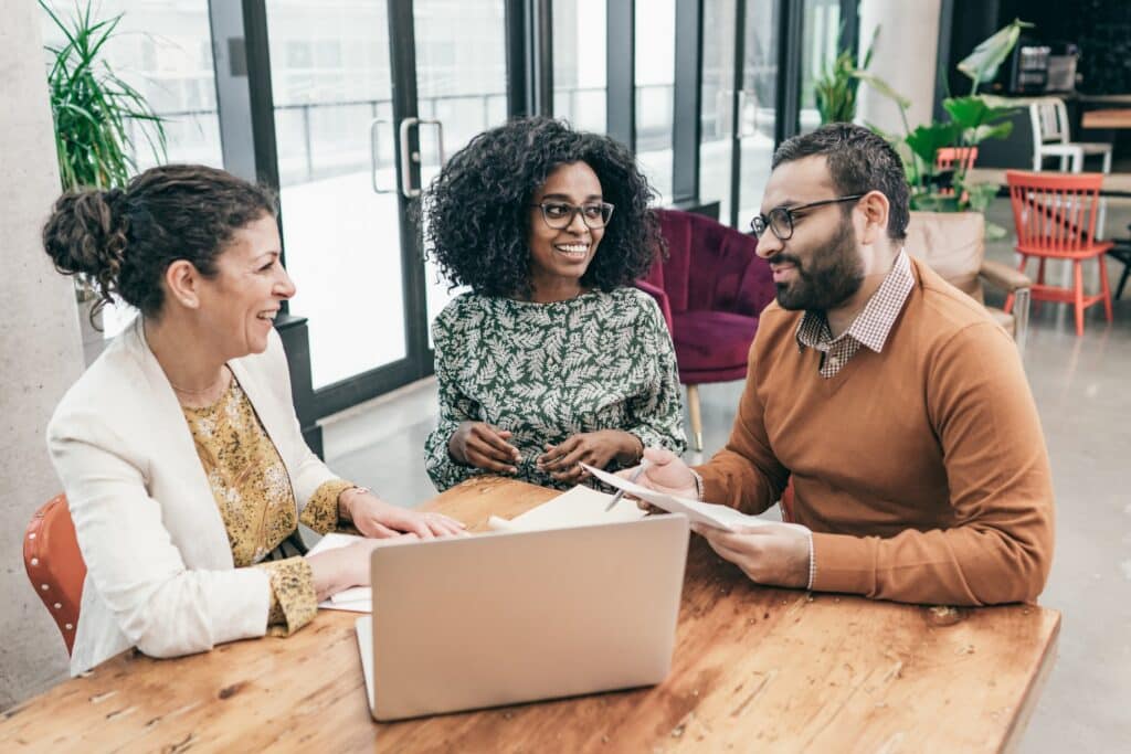 Three smiling professionals sitting around a wooden table in a modern office, collaborating over documents and a laptop.