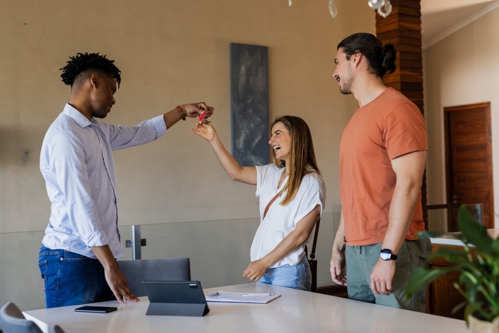 A real estate agent handing a set of keys across a table to an excited young couple in a modern home.