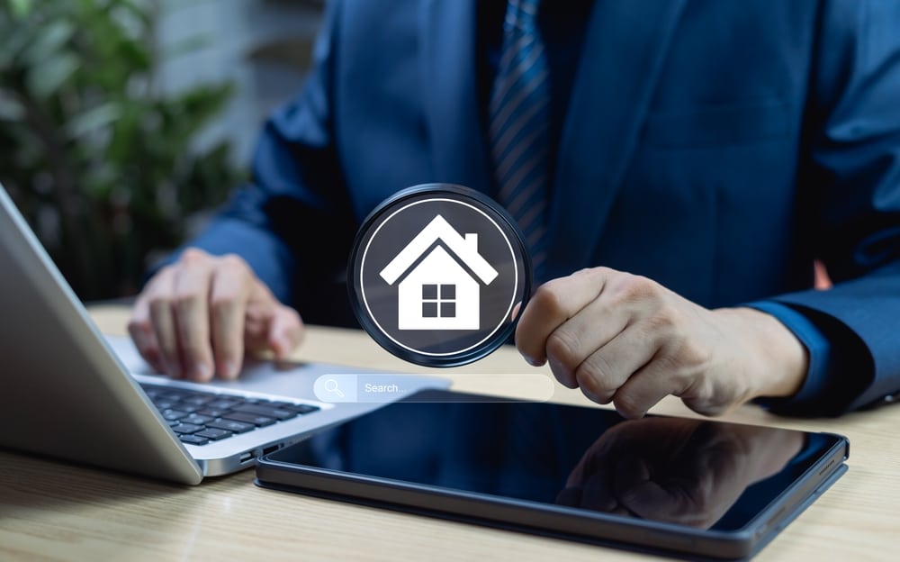 A person in a blue business suit working on a laptop, featuring a digital overlay of a magnifying glass containing a house icon above a search bar.